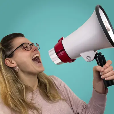 Eine Frau mit langen, blonden Haaren und Brille hält ein Megafon in der Hand und spricht laut hinein. Sie trägt einen rosa Pullover und lächelt leicht, während sie ihre Botschaft durch das Megafon verbreitet. Der Hintergrund ist einfarbig in einem hellen Türkis gehalten.