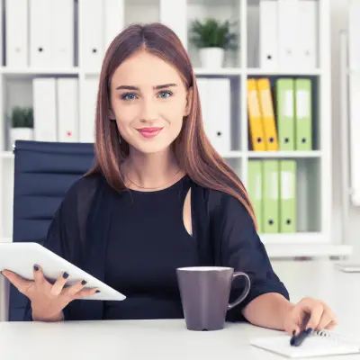 Eine junge Personaldienstleistungskauffrau, die im Bereich der Personalbeschaffung tätig ist, sitzt lächelnd in ihrem Büro. Sie hält ein Tablet in der linken Hand und schreibt mit der rechten Hand in ein Notizbuch. Vor ihr steht eine Tasse auf dem Schreibtisch. Im Hintergrund sind Regale mit Ordnern und Büromaterialien zu sehen.