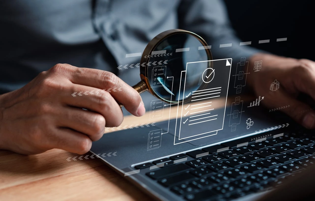 Zwei Hände halten eine Lupe über einen Laptop auf einem Holztisch. Über dem Laptop schweben digitale holografische Grafiken: Dokumente mit Häkchen und Checklistenpunkten, vernetzte Workflow-Knoten, ein Balkendiagramm und Datenverarbeitungs-Icons – symbolisch für Dokumentenprüfung, Audits oder digitale Analyse.